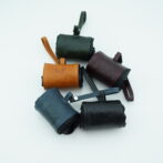 Group of small leather pouches in orange, green, burgundy, blue, and black with wrist straps and stitched edges on a light background.