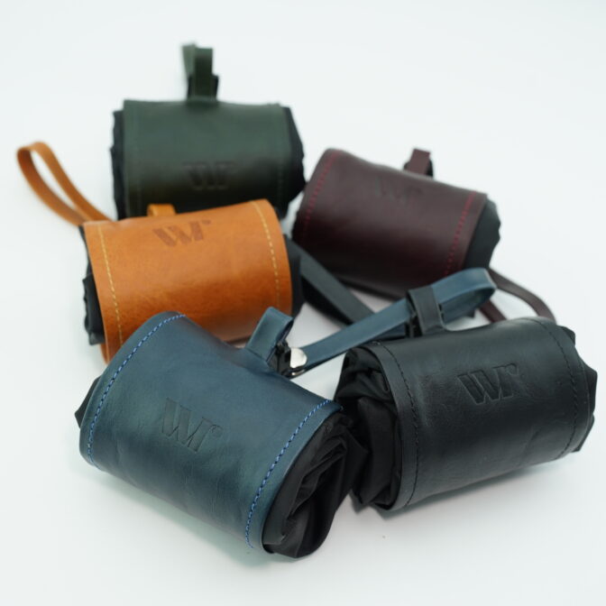 Pile of small leather pouches in blue, tan, green, and burgundy stacked together, showing stitched edges and straps.