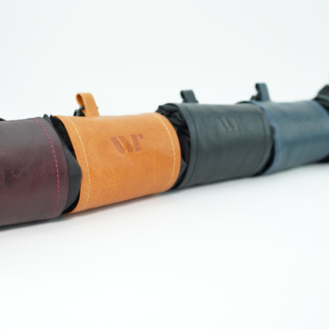 Row of leather camera or tool pouches in burgundy, orange, navy, and green, showing stitched edges and embossed 'W' logo.