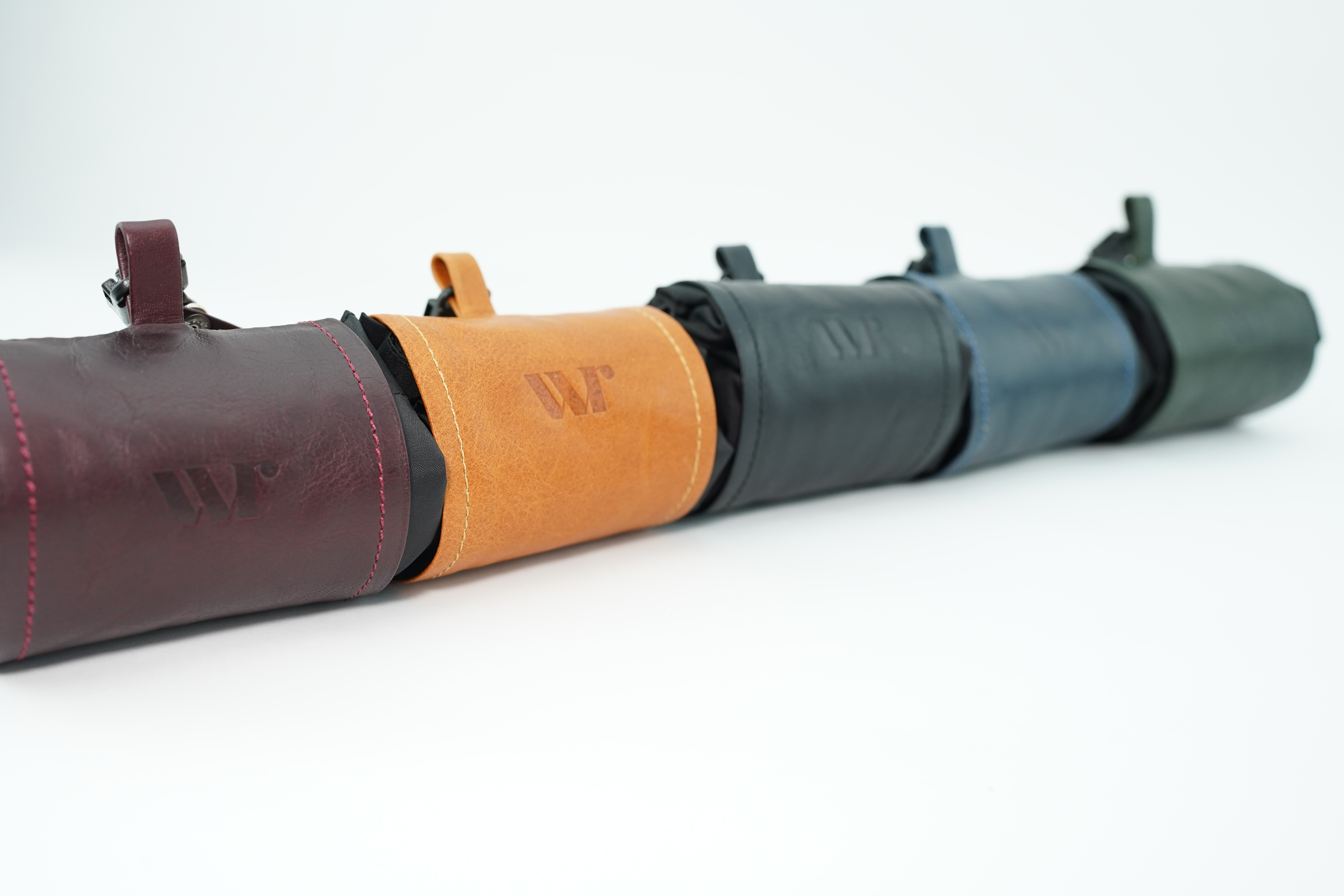 Row of leather camera or tool pouches in burgundy, orange, navy, and green, showing stitched edges and embossed 'W' logo.