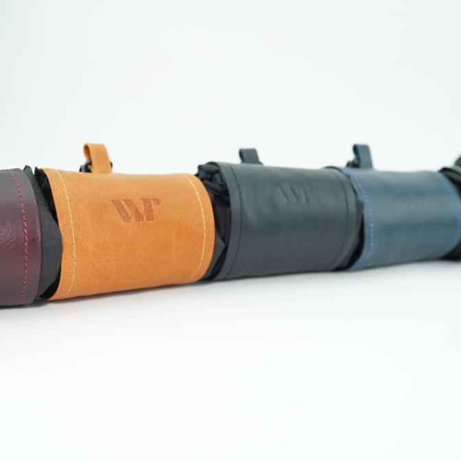 Line of colorful leather pouches arranged in a row on a white surface (burgundy, orange, gray, blue, green).