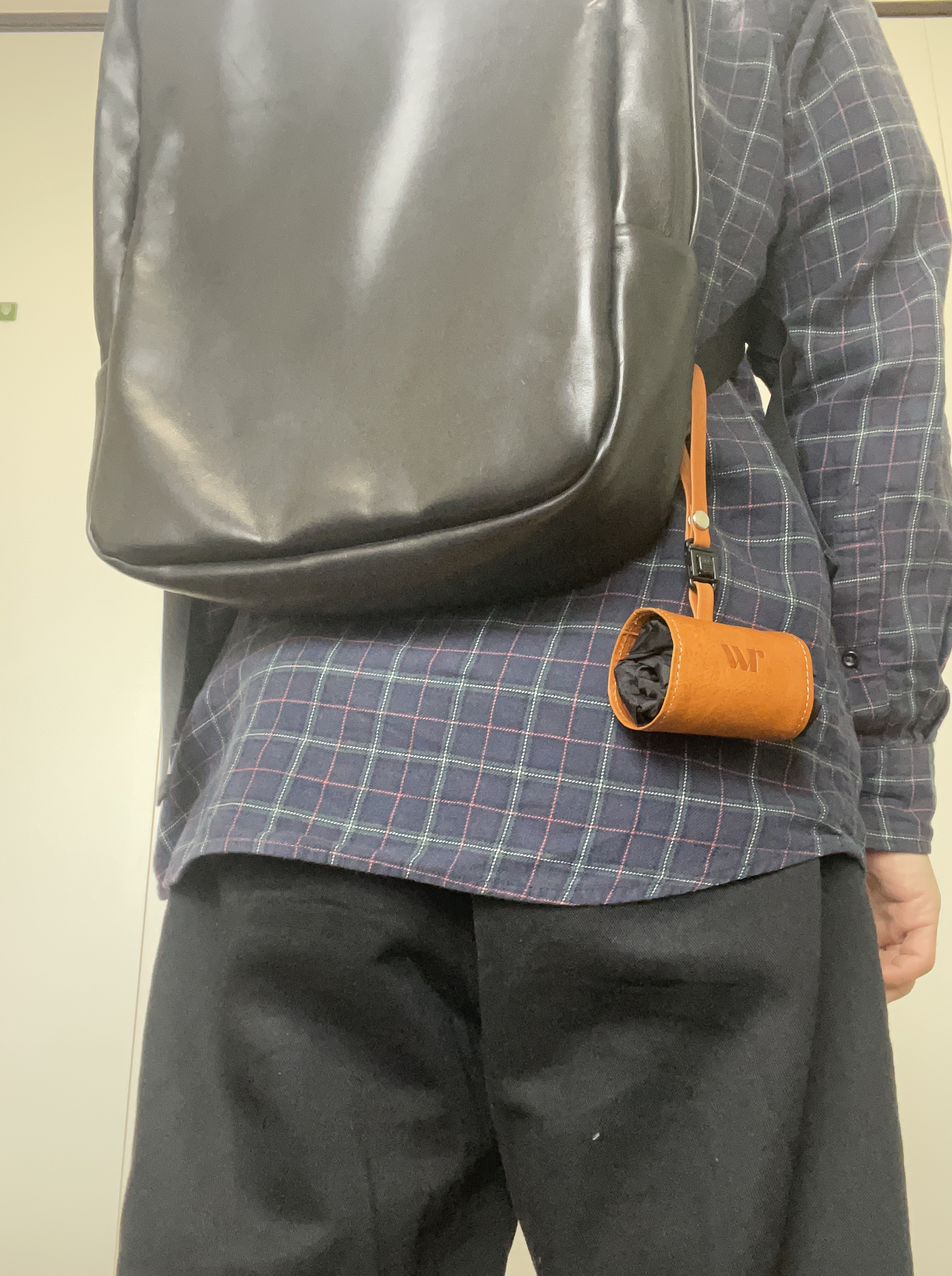 A person wearing a large black leather backpack over a dark plaid shirt with brown leather strap accessories visible to the side.