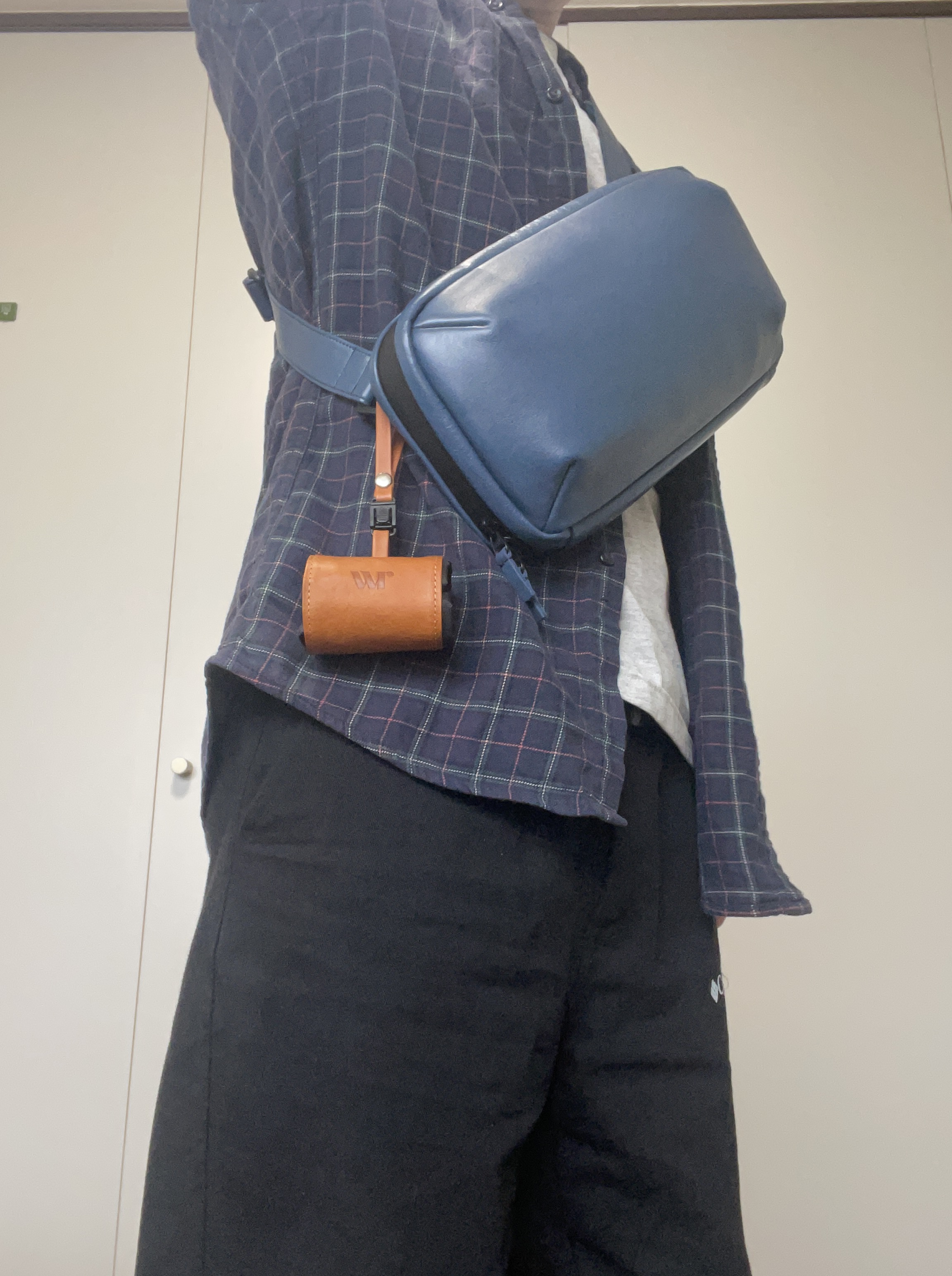 Person in a blue plaid shirt seen from the side with a blue crossbody bag strapped around the waist and a small brown leather pouch hanging from the strap.