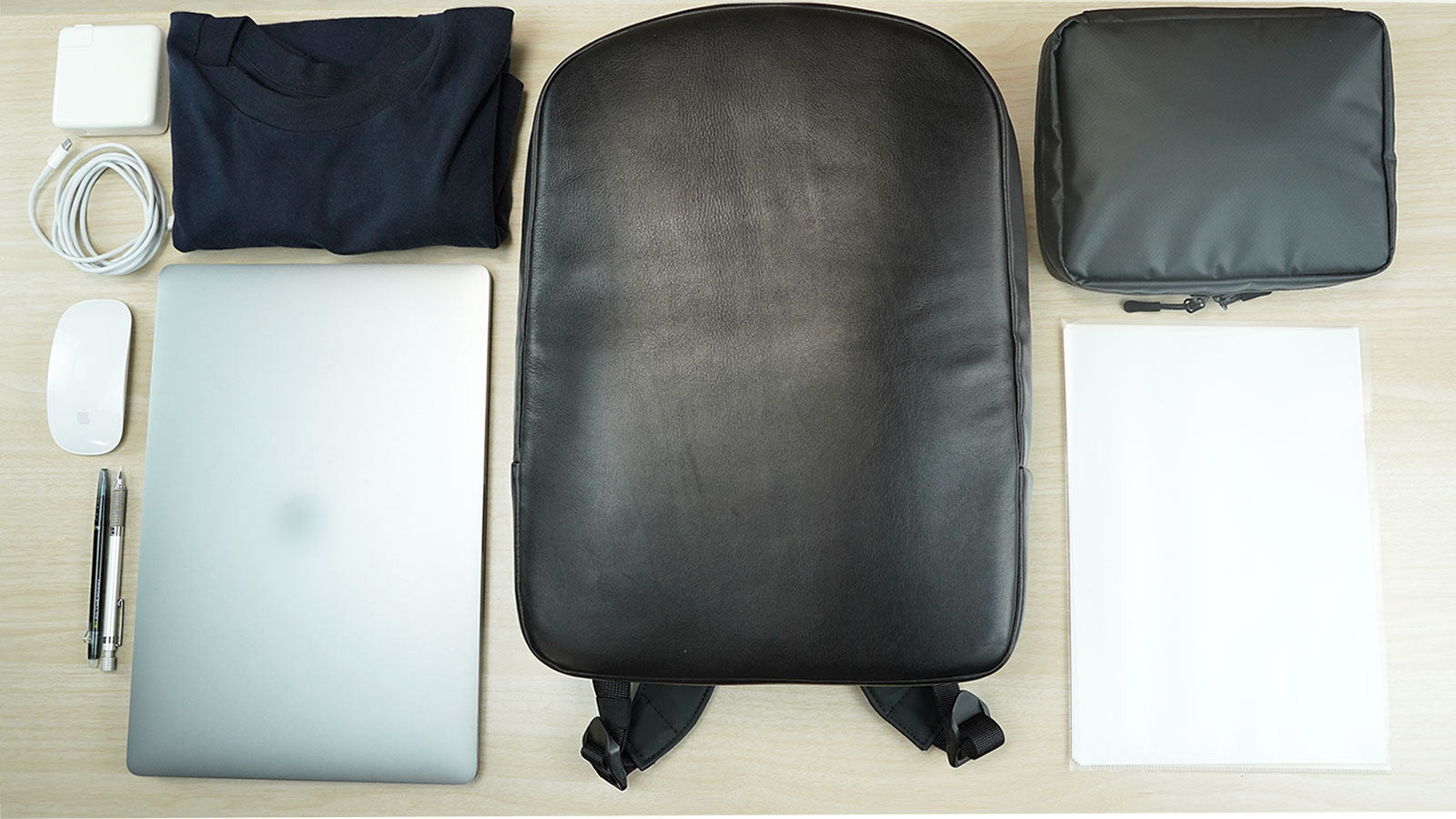 Flat lay: black backpack centered on a wooden desk, surrounded by a laptop, mouse, charger, t‑shirt, pens, pouch, and notepad.